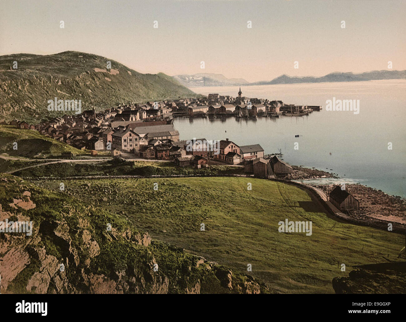 Una stampa fotocromatica di Hammerfest, una città situata a Finnmark, Norvegia, catturata dal nord. L'immagine mostra l'ambientazione costiera della città, circondata da montagne e acqua, evidenziando il paesaggio unico della Norvegia settentrionale e il significato storico della città. Foto Stock