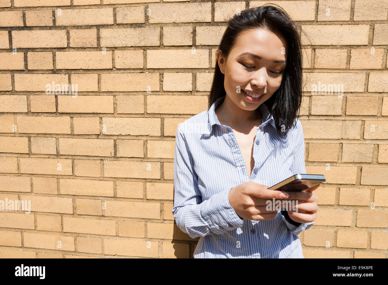 Sorridente giovane imprenditrice di messaggistica di testo sul telefono cellulare contro un muro di mattoni Foto Stock