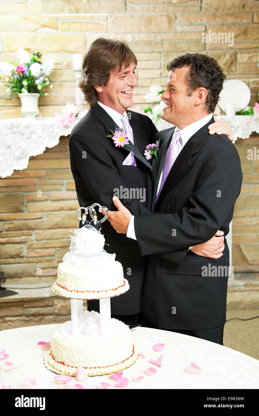 Due bello uomini gay in tuxedos abbracciare con amore al loro ricevimento di nozze. Torta di Nozze e petali di rosa in primo piano. Foto Stock