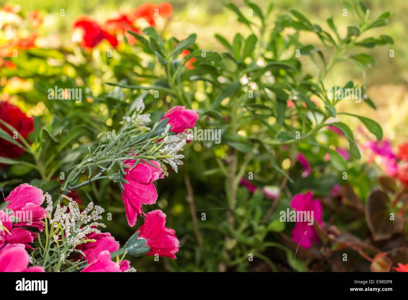 Plastica e reale fiori votive sotto un albero di supporto di un edicola votiva nella regione Emilia Romagna in Italia settentrionale. Semplice paese fiori sono ai piedi della struttura circondata dal verde di erbe infestanti: i colori sono giallo, rosa fucsia, viola, bianco Foto Stock