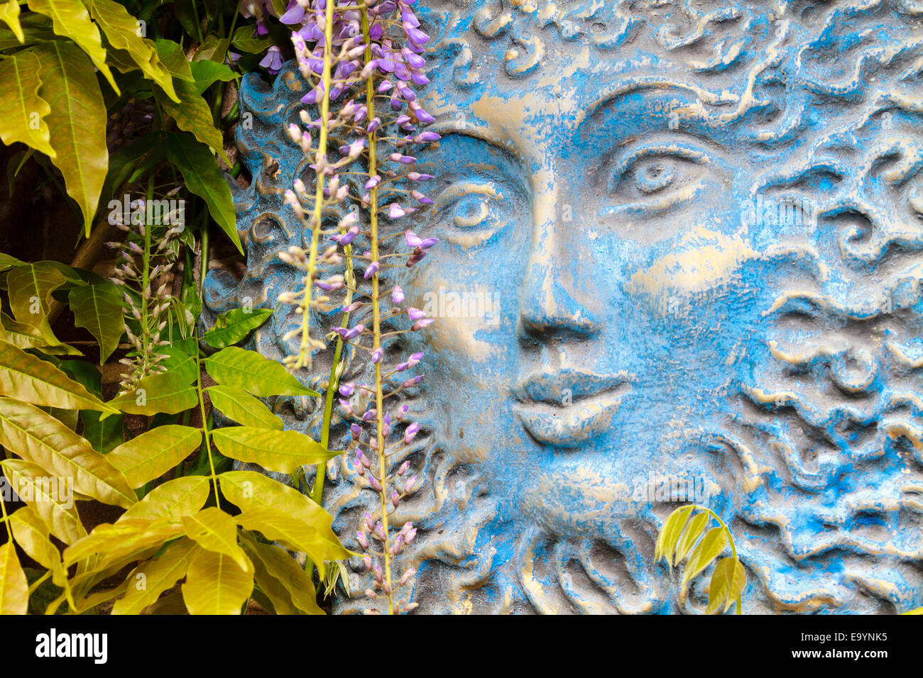 Big Blue sun faccia appeso a una parete del giardino dietro foglie pendenti Foto Stock