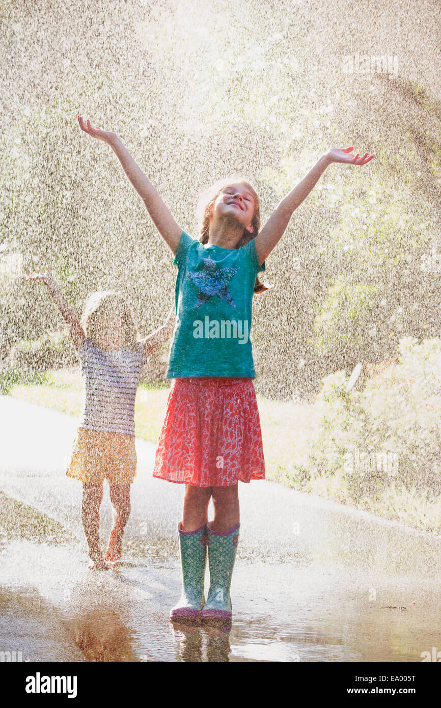 Due ragazze con i bracci aperti in piedi in uno spruzzo di acqua su strada Foto Stock