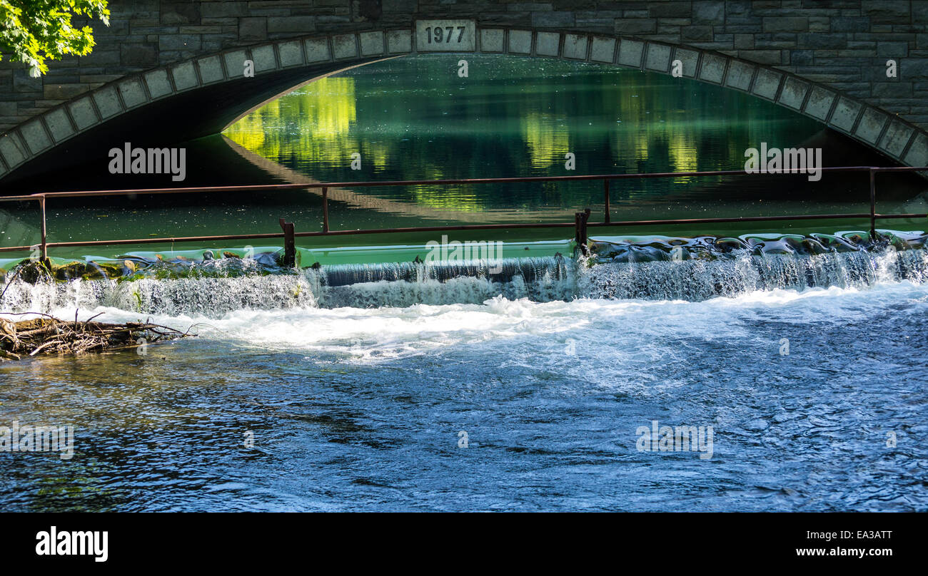 La calma acqua riflette la luce del sole sotto il ponte ad arco con una piccola cascata in primo piano. Foto Stock