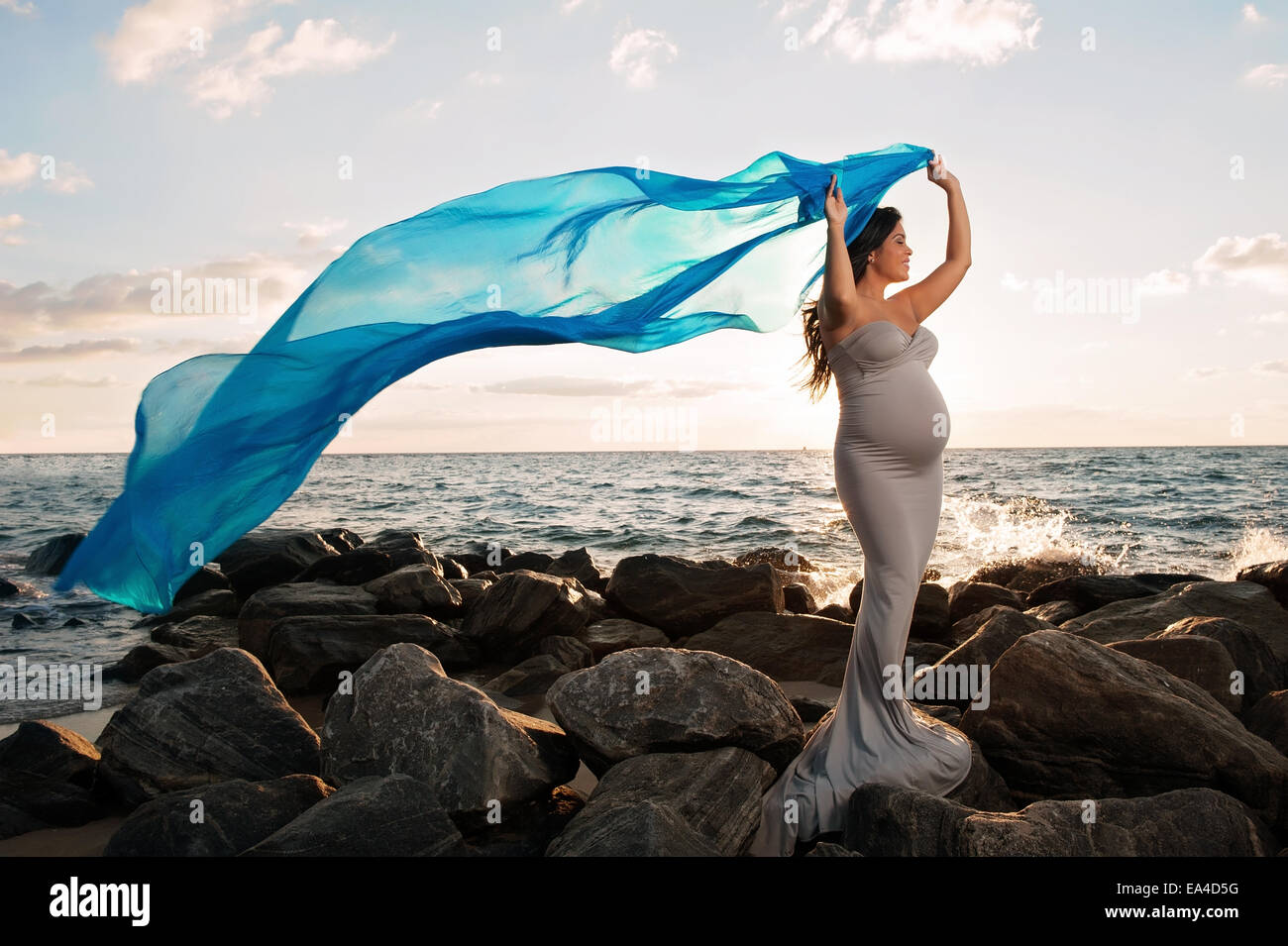 Una bella e sorridente donna incinta su una spiaggia rocciosa di sunrise. Lei è in possesso di un blu velo di seta che scorre nel vento. Foto Stock