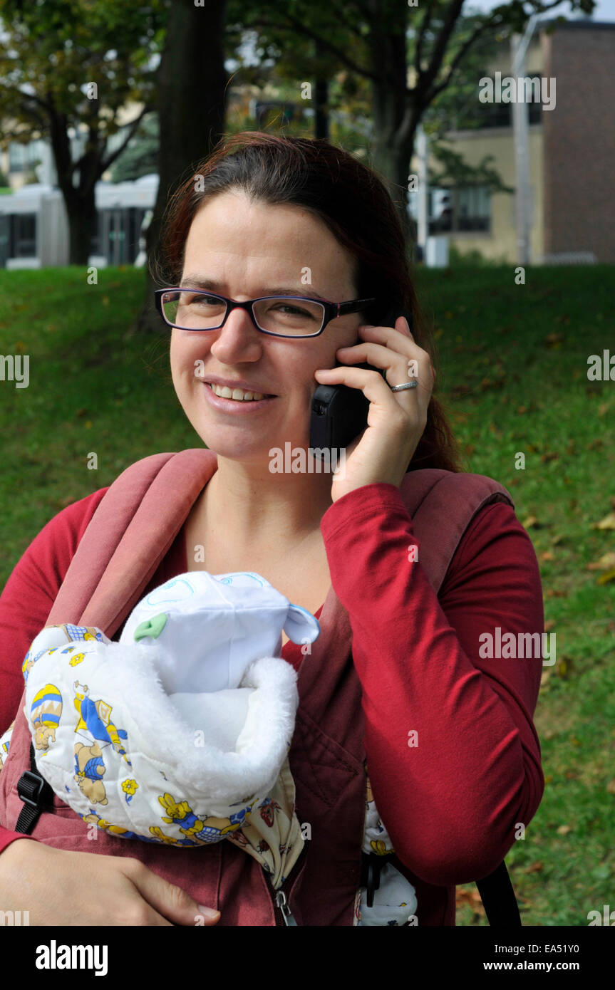 Un sorridente madre che porta un bambino e utilizzando un telefono cellulare Foto Stock