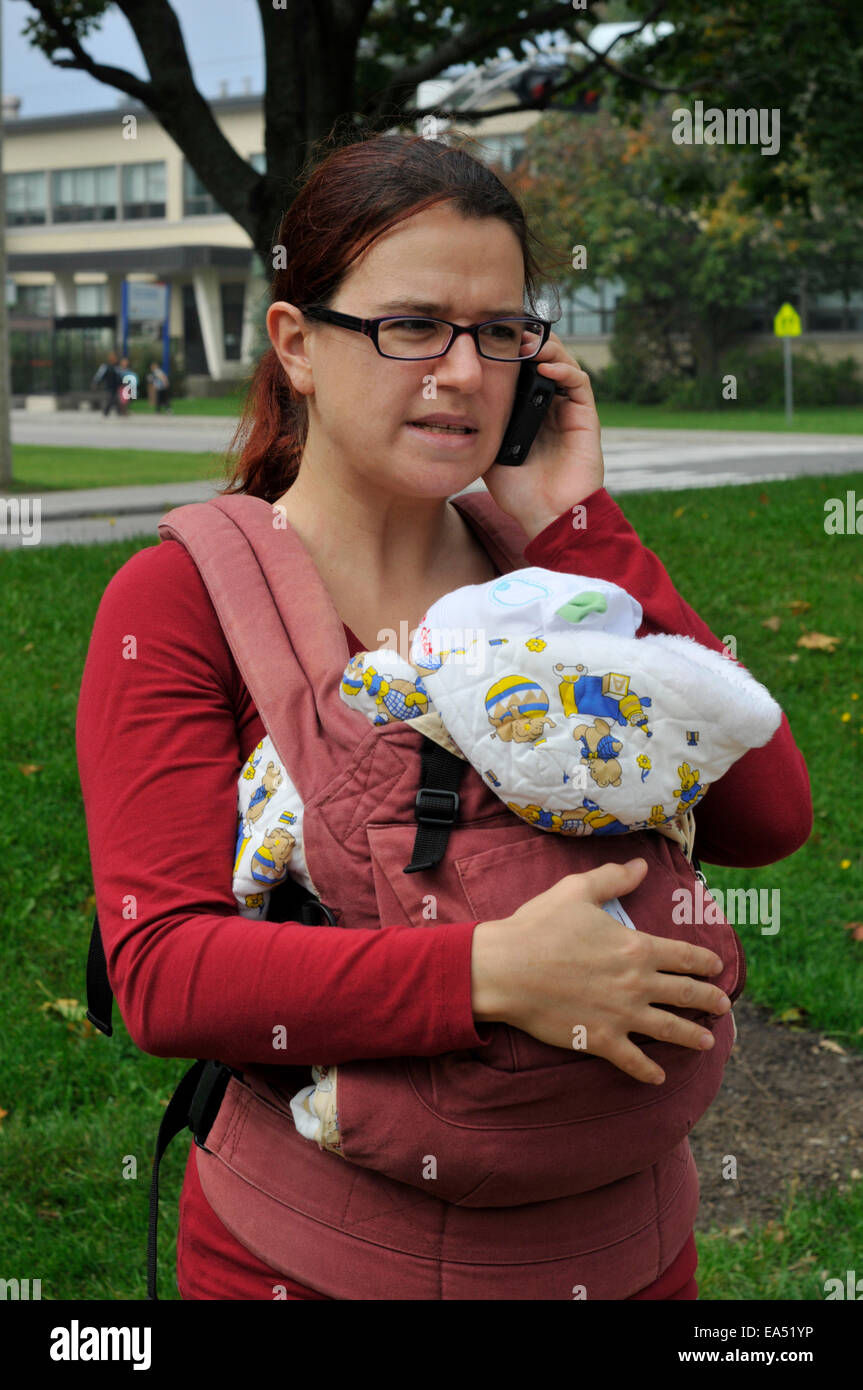 Una madre che porta un bambino e utilizzando un telefono cellulare, con un volto arrabbiato Foto Stock