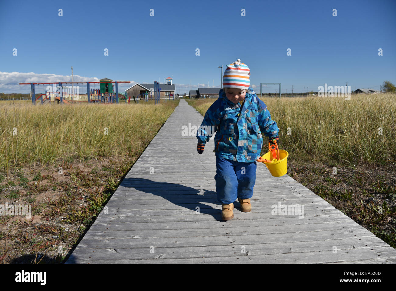 Un ragazzino a piedi lungo una passerella di legno con un cucchiaio e spade di andare in spiaggia. Il suo autunno in modo che egli è in un cappotto e cappello Foto Stock