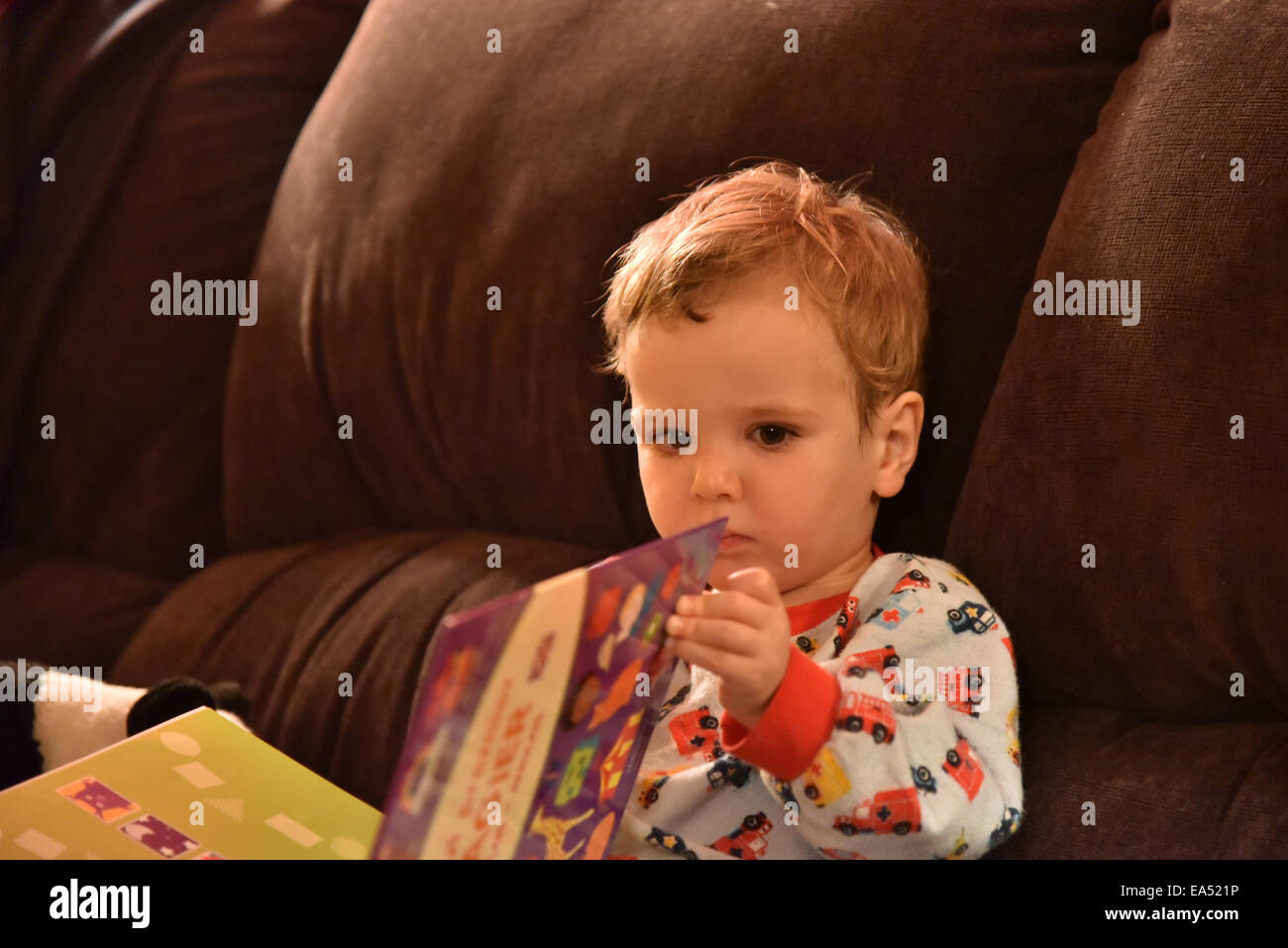 Un giovane ragazzo (due e una metà di anni) in pigiama seduto su un divano a leggere un libro appena prima di andare a dormire Foto Stock
