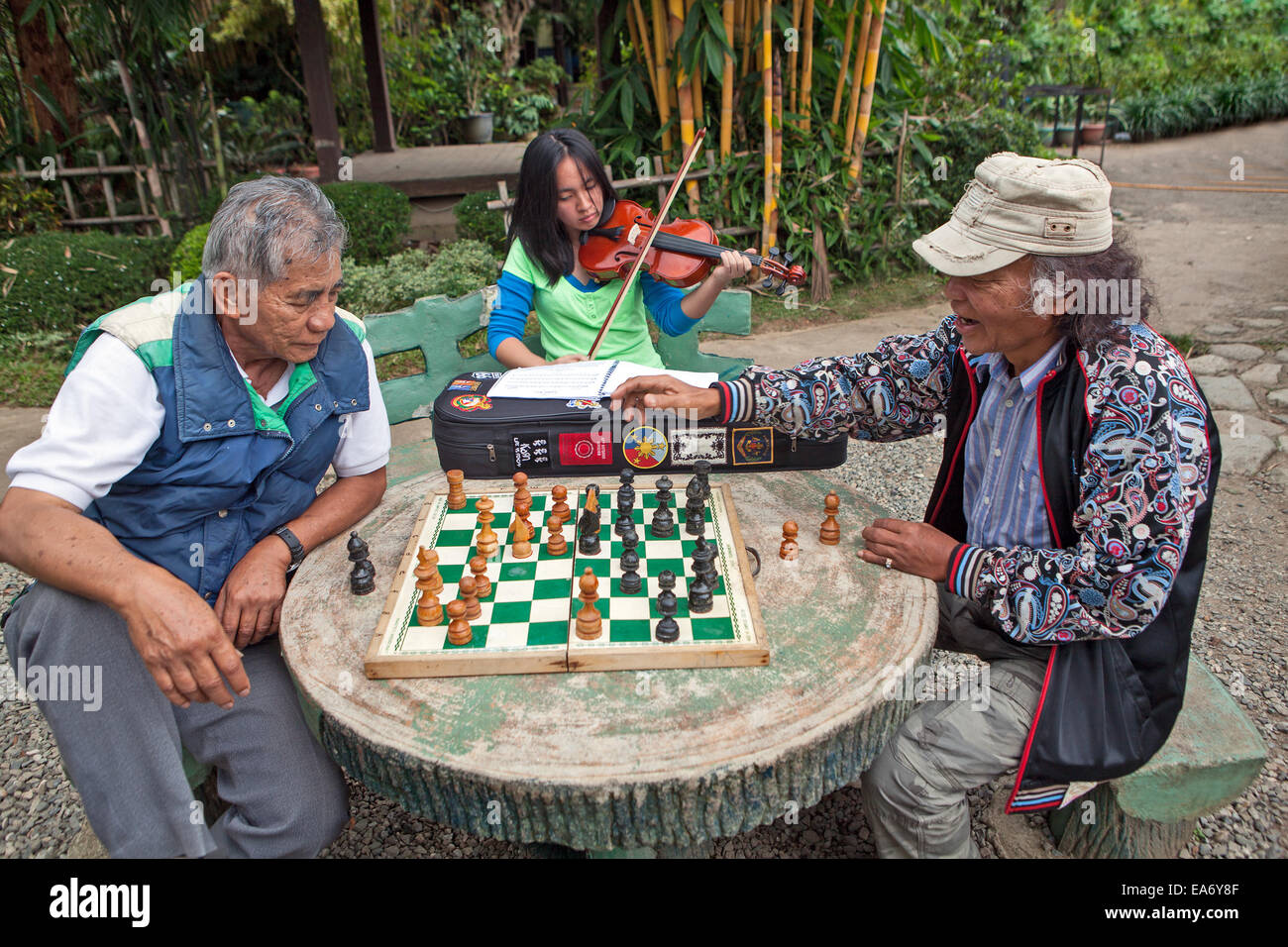 Due uomini filippini giocare a scacchi mentre una giovane ragazza svolge il suo violino a Burnham Park, Baguio City nelle Filippine. Foto Stock