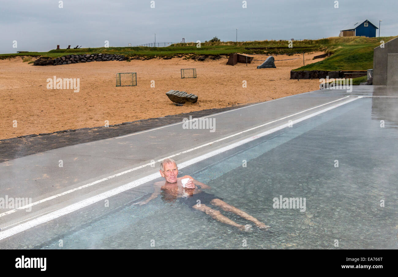 Uomo locale si riscalda se stesso con una tazza di tè in un materiale termicamente piscina riscaldata a Ylstrond, Reykjavik è solo la spiaggia della sabbia Foto Stock