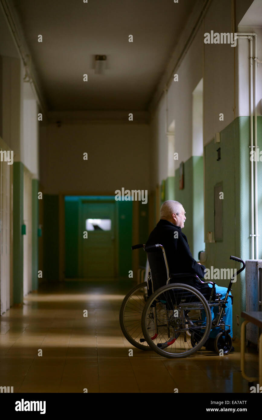 Il vecchio uomo su una sedia a rotelle guardando fuori da una finestra da sola su un lungo corridoio Foto Stock