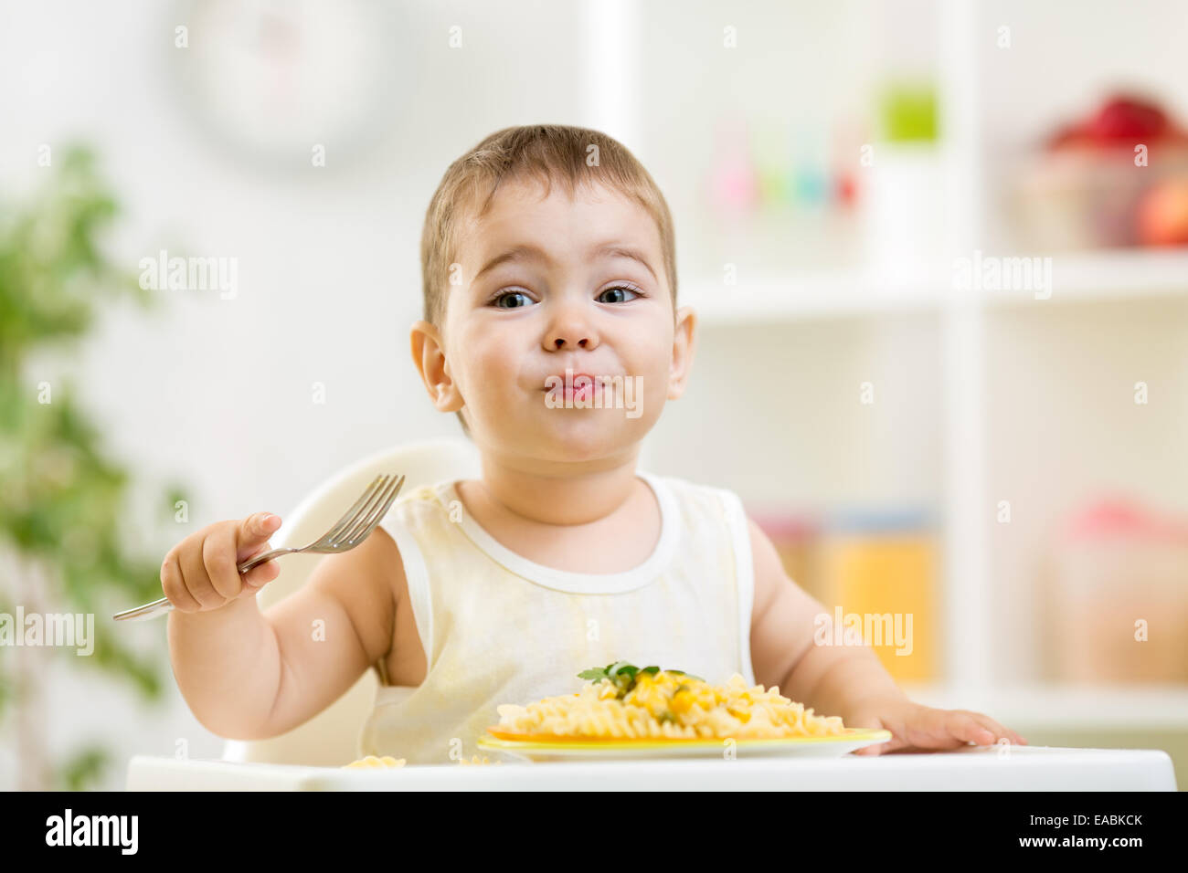 Carino kid ragazzo in un seggiolone per alimentare con una forcella e una pasta Foto Stock