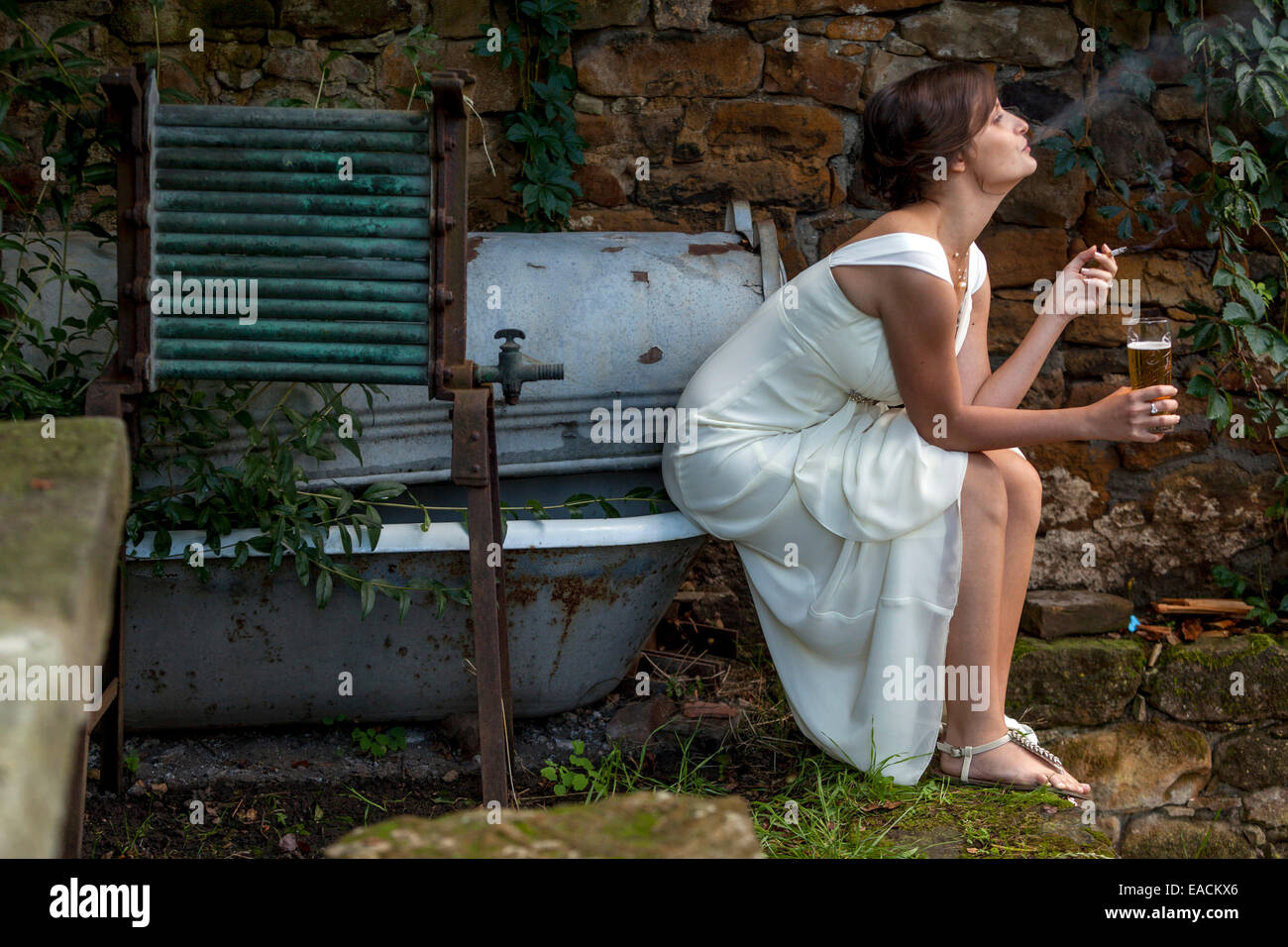 Sposa in abito bianco bevendo birra donna che fuma sigaretta riposante da sola in un angolo tranquillo del Garden Outdoor Scene Wedding Break Sposa che fuma sigaretta Foto Stock