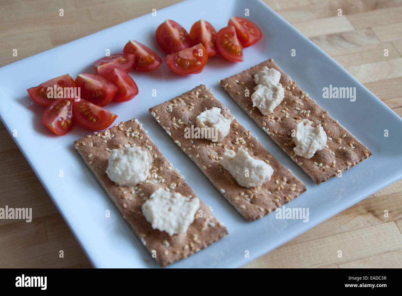Semplice snack di ak-mak crackers, humus e fette di pomodori ciliegia su una piastra quadrata. Foto Stock