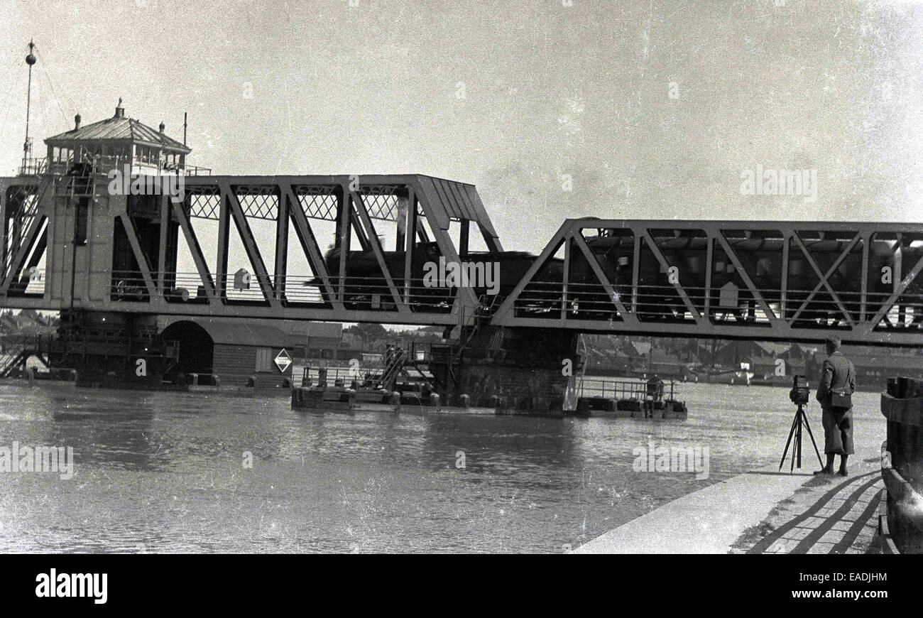 1930s, foto storiche che mostra un fotografo utilizzando una videocamera e treppiede per scattare una foto di un treno che attraversa un fiume. Foto Stock