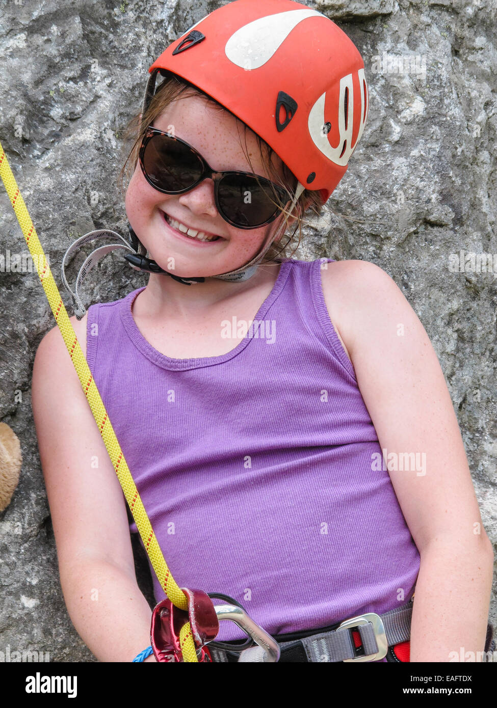 Sorridente ragazza giovane con occhiali da sole e arrampicata attrezzature comprensiva di casco e il sistema di cavi in attesa di salire una parete di roccia. Foto Stock