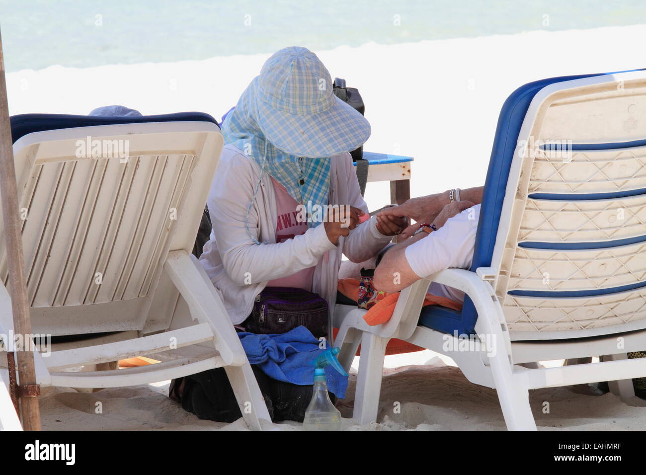 Thai donna che indossa hat rendendo unghie Manicure pedicure per un uomo sulla chaise longue (letto da giorno) sulla spiaggia. Pattaya, Thailandia Foto Stock