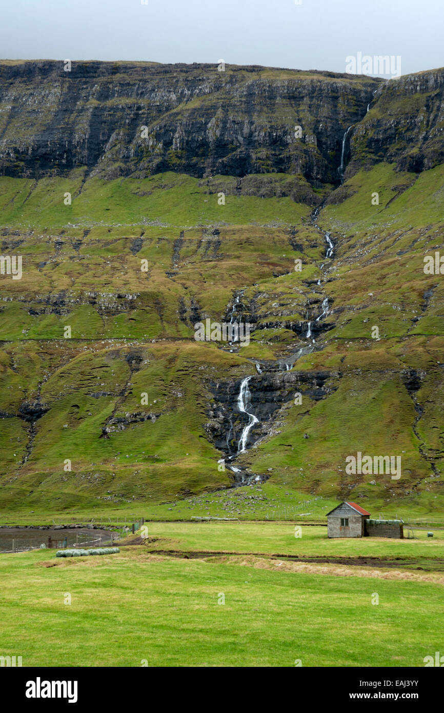 Una piccola casa sotto le ripide pendici di una collina verde con cascata, isole Faerøer Foto Stock