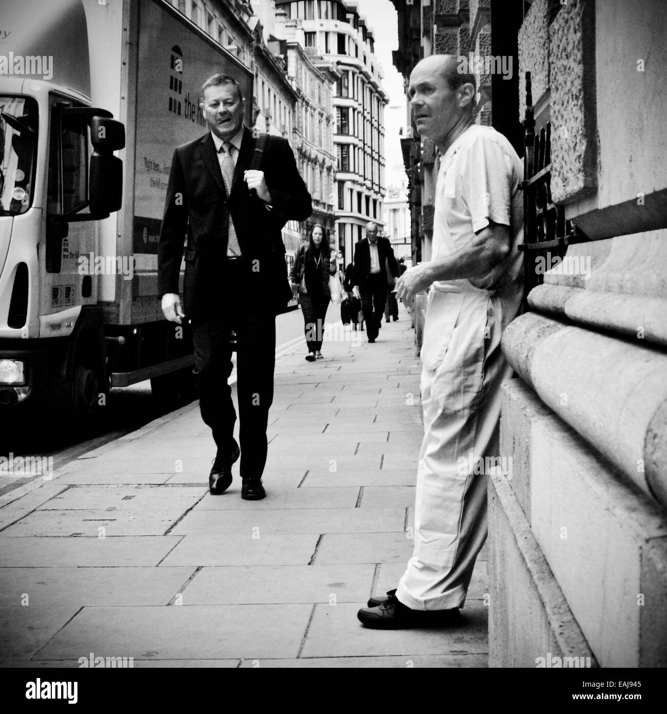 Un tuttofare prende una pausa sigaretta come un lavoratore cittadino passeggiate passato lui a Londra Foto Stock