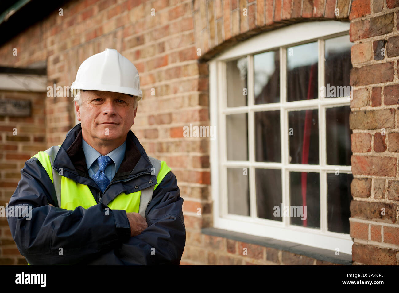 Ispettore edificio, con le braccia piegate, in piedi accanto alla finestra di un edificio in mattoni. Foto Stock