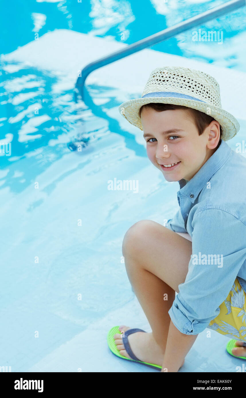 Ritratto di ragazzo sorridente indossando cappello di paglia accovacciato da piscina Foto Stock