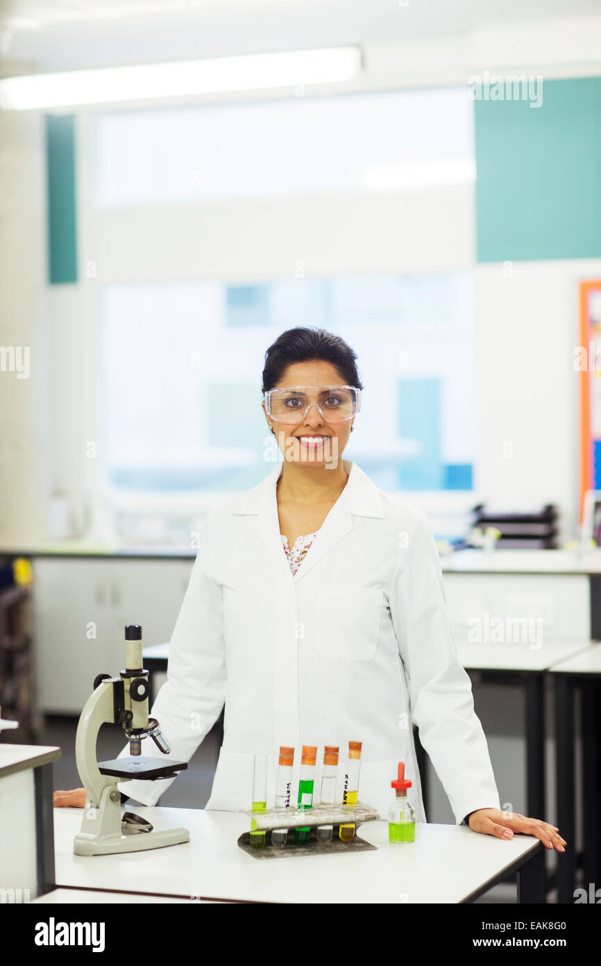 Ritratto di donna sorridente insegnante indossando occhiali protettivi, in piedi dietro la scrivania con microscopio e provette per la prova in rack Foto Stock