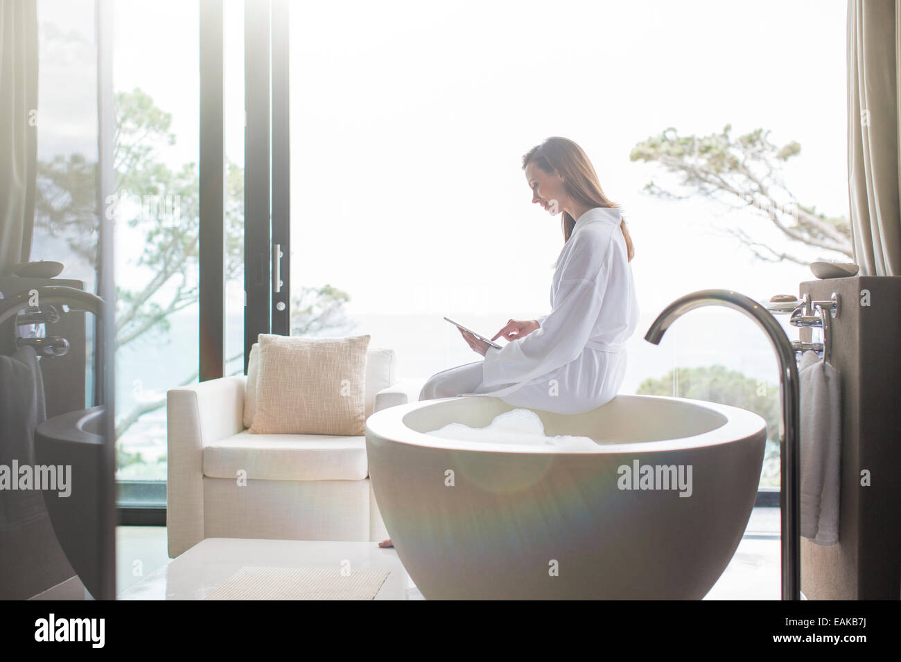 Donna che indossa accappatoio bianco seduto sul bordo della vasca da bagno moderna e con tavoletta digitale Foto Stock