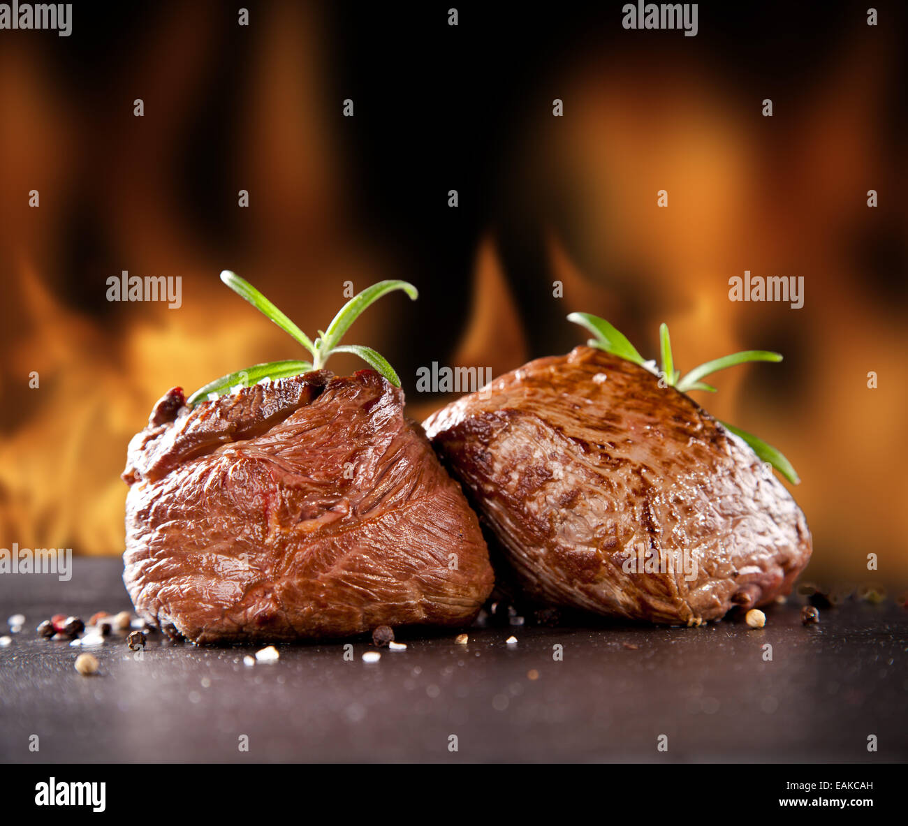 Pezzi di carne rossa bistecche con rosmarino servita sulla pietra nera superficie. Blur fiamme di fuoco sullo sfondo Foto Stock