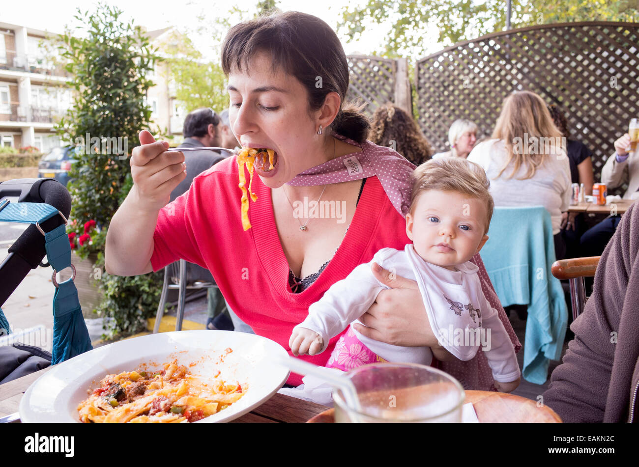 Madre mangiare pasta al ristorante e azienda piccola tre mese bambina tra le sue braccia, London, England, Regno Unito Foto Stock