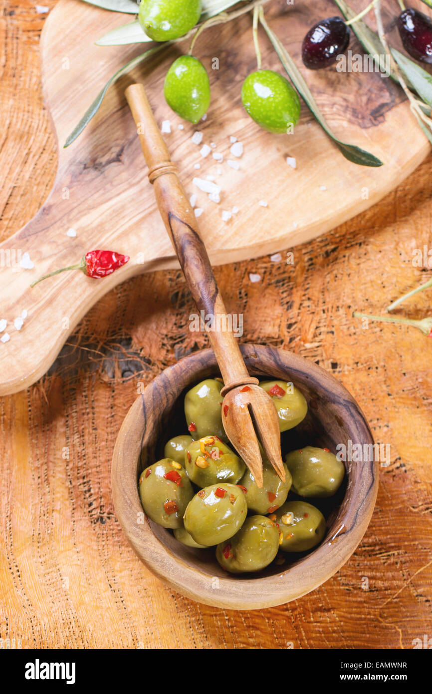 Olive verdi in legno d'olivo ciotola con peperoncino e olio d'oliva di filiali su un tavolo di legno. Foto Stock