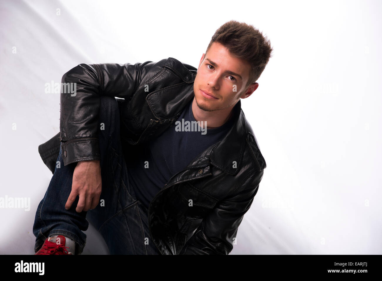 Bel giovane uomo o ragazzo seduto sul pavimento bianco, vestita di nero giacca di pelle Foto Stock