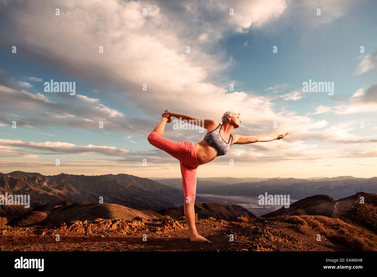 Senior donna fare yoga pone sulla cima della montagna al tramonto con il paesaggio deserto e la valle sottostante. Foto Stock