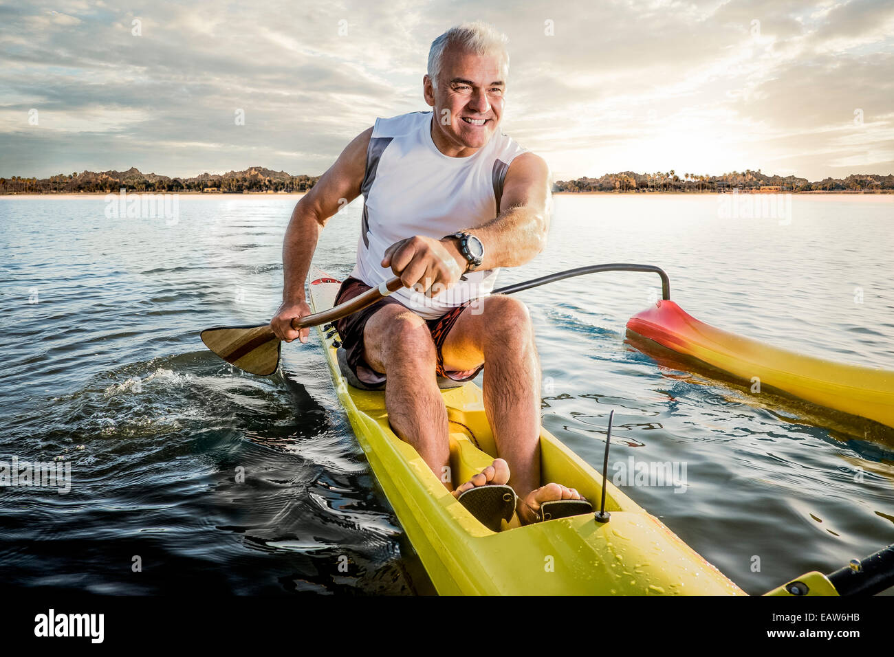 Uomo Senior canoa outrigger canoe nell'oceano al tramonto con isola tropicale dietro di lui. Foto Stock