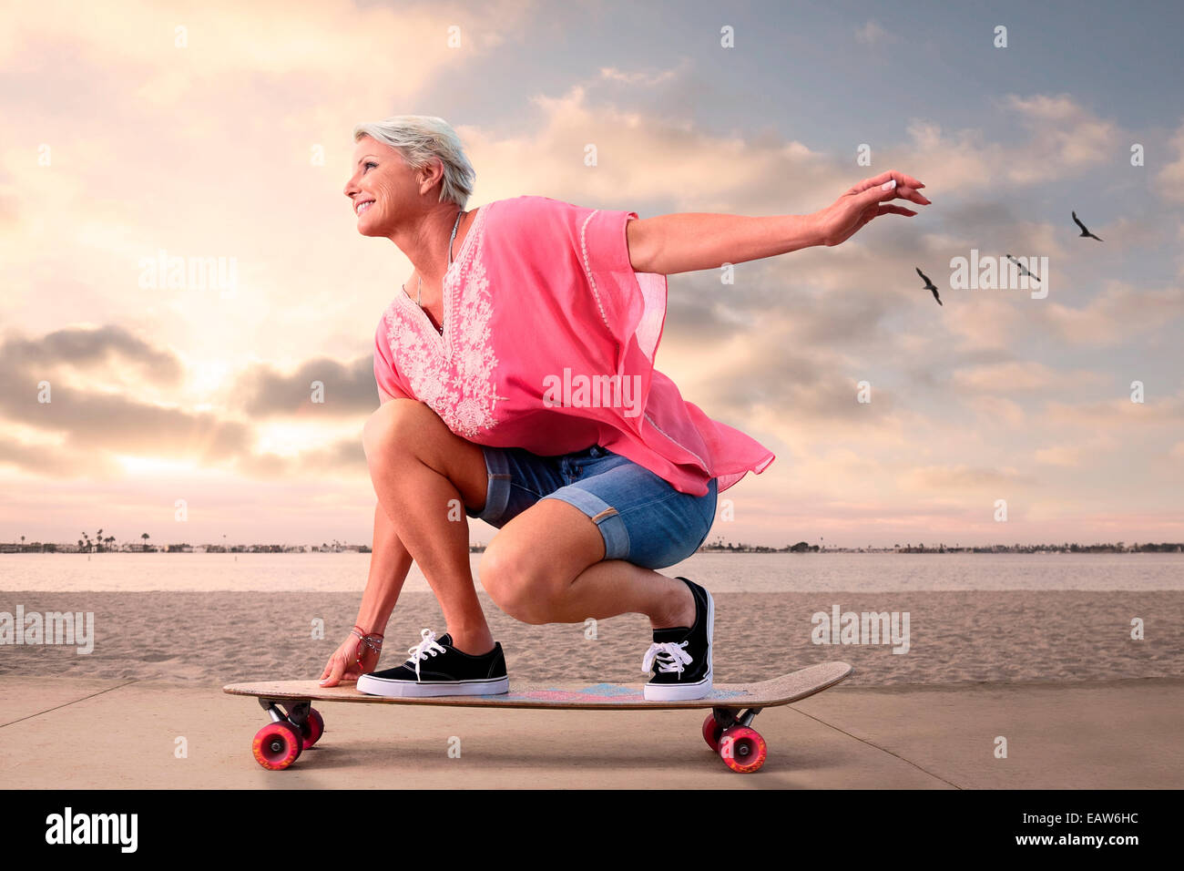 Giovane donna senior su skateboard lungo la spiaggia al tramonto, uccelli in volo in background. Foto Stock