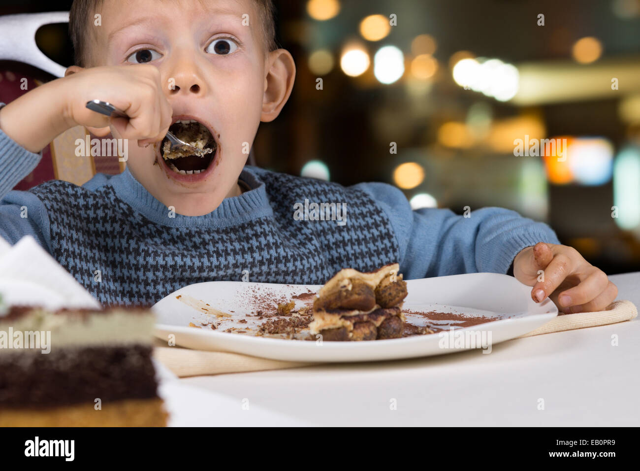 Fame little boy gobbling giù una fetta di gustosa torta al cioccolato con la bocca spalancata per un boccone come egli si siede a un tavolo in un ristorante. Foto Stock