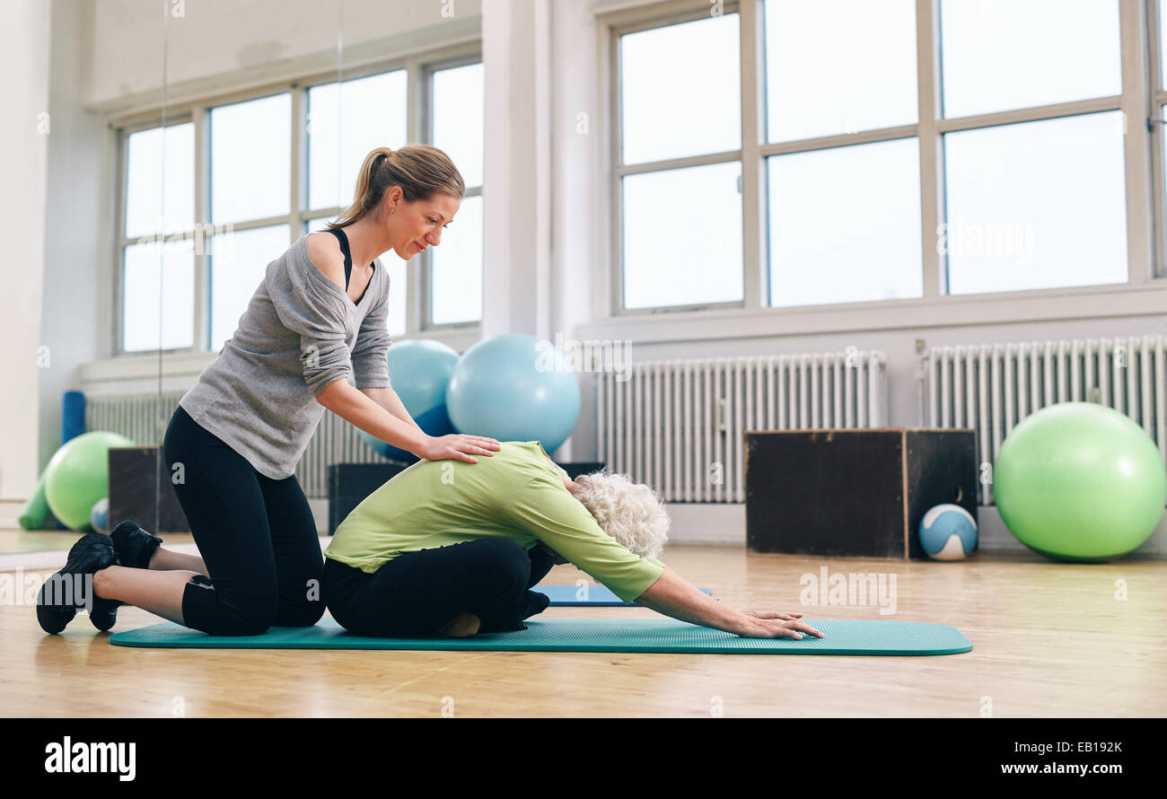 Trainer femminile aiutando donna senior di chinarsi. Vecchia donna fare yoga su un tappeto di esercizio con terapista fisico contribuendo alla palestra. Foto Stock