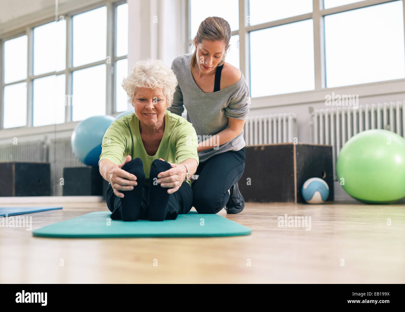 Donna anziana essendo aiutata dal suo istruttore in palestra per esercizio. Senior donna seduta sul tappetino fitness la piegatura in avanti e Foto Stock