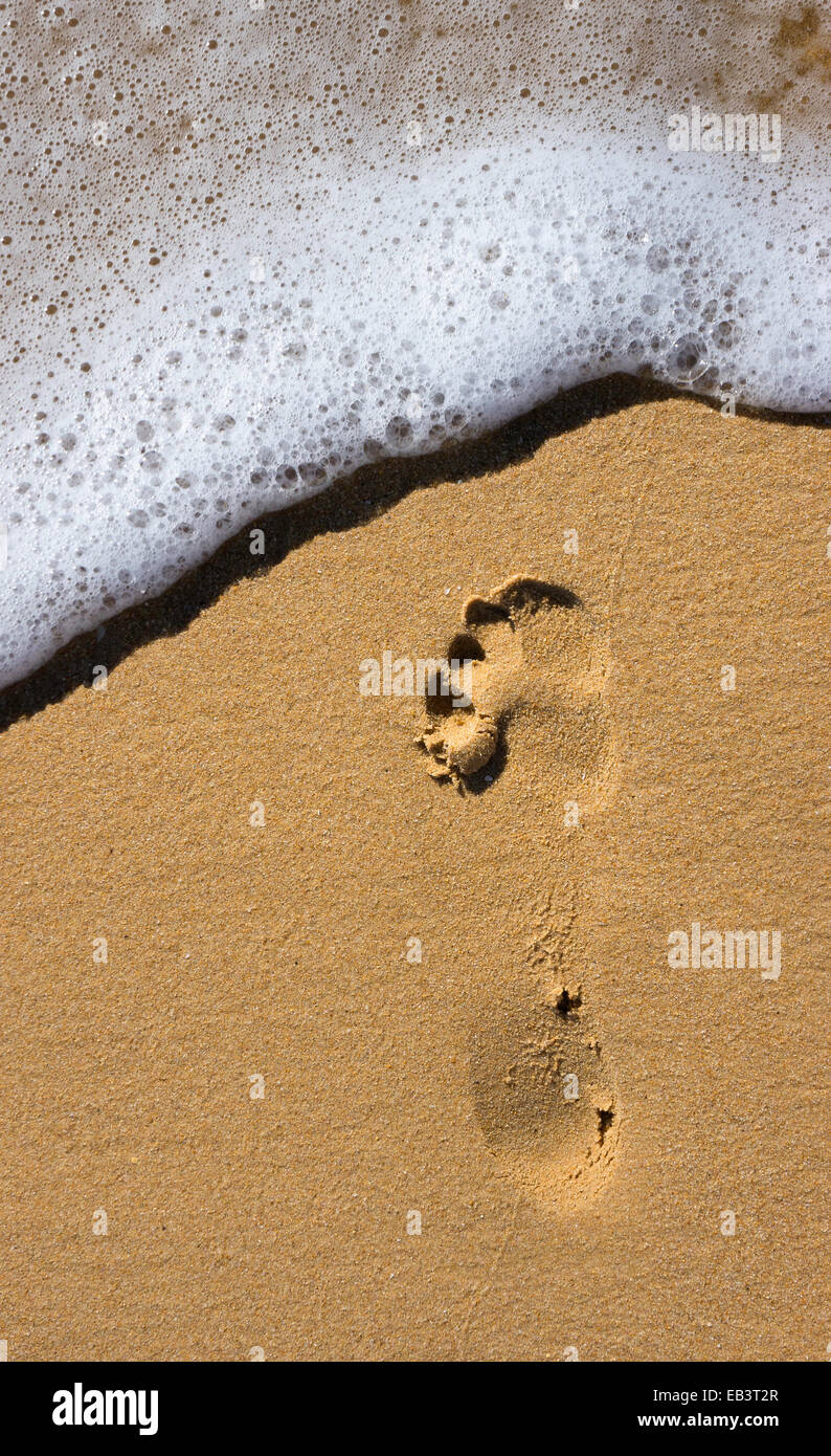 Una singola impronta su di una spiaggia soleggiata con un onda di schiumatura circa per lavare oltre l'impronta del piede. Foto Stock