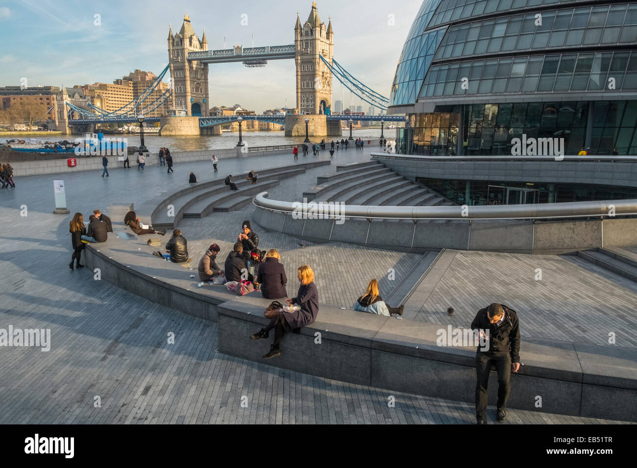 Le persone al di fuori seduta rilassante vicino al municipio edificio, con Landmark Tower Bridge in background, London, England Regno Unito Foto Stock