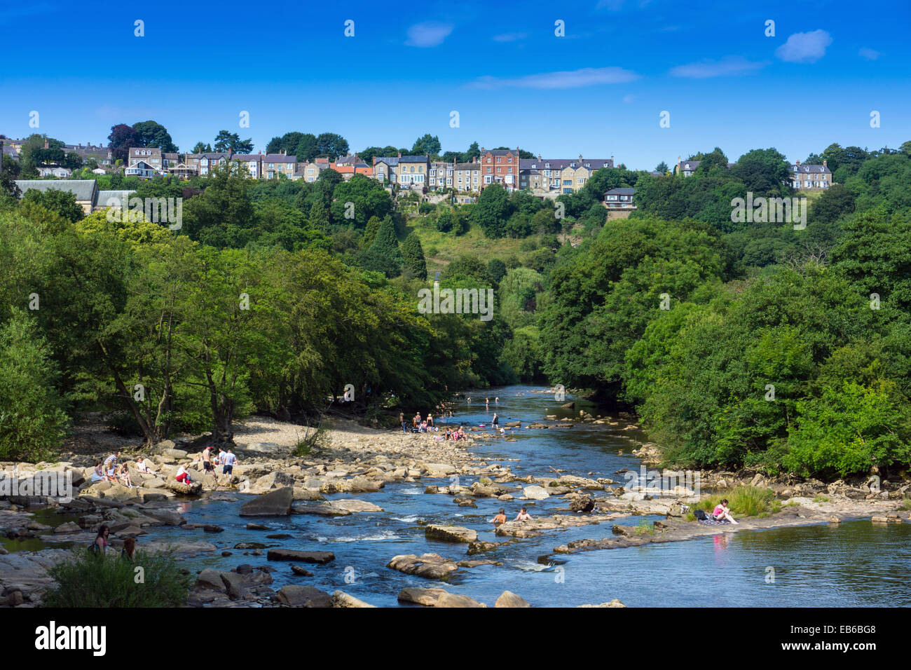Le persone che giocano in acqua, estate sul fiume Swale, con alberi verdi e la linea di case al di sopra di Richmond, North Yorkshire, Inghilterra Foto Stock