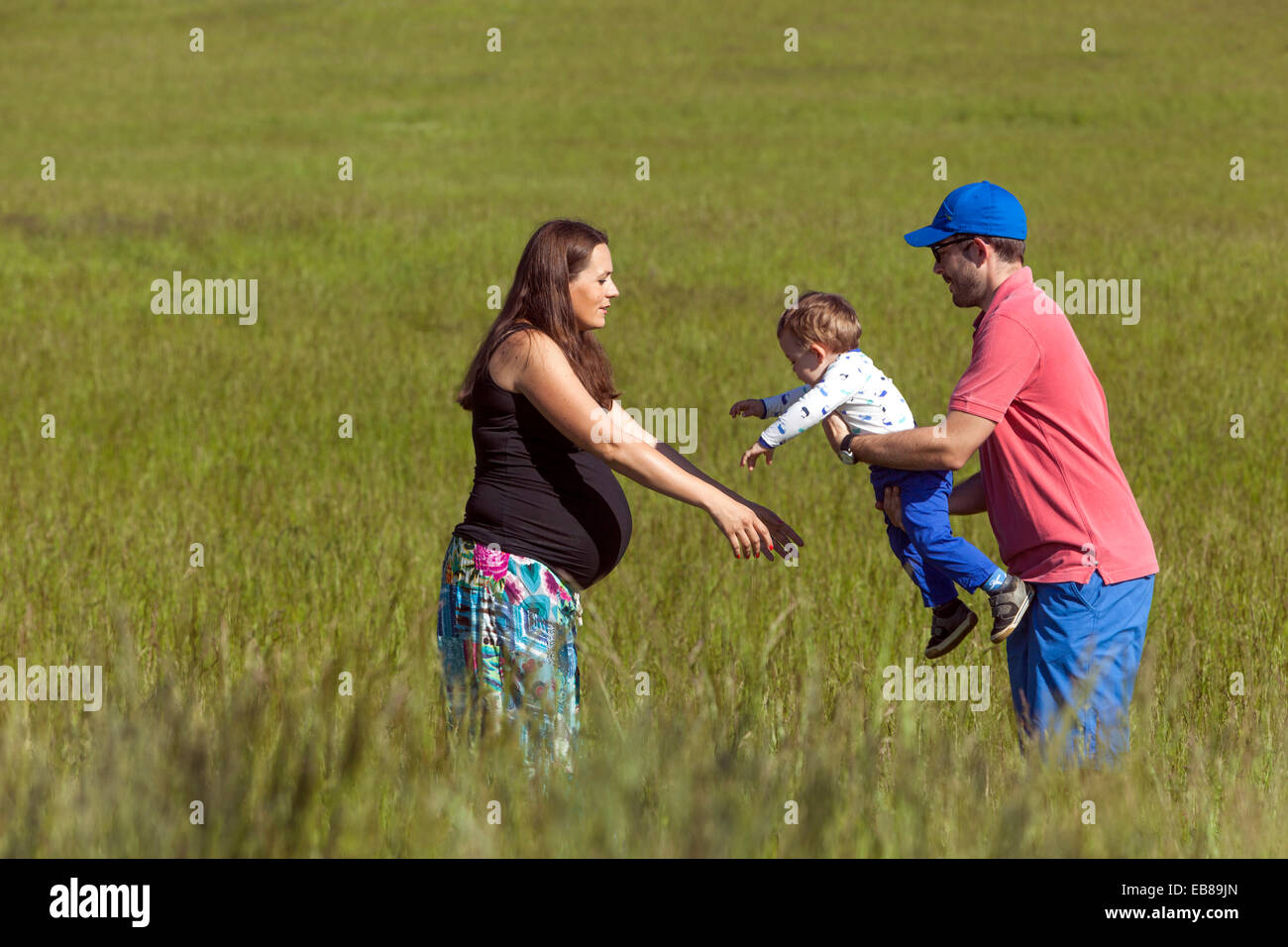 Una famiglia felice, una donna incinta con il marito e il bambino trascorrere del tempo insieme e giocare in un prato estivo Foto Stock
