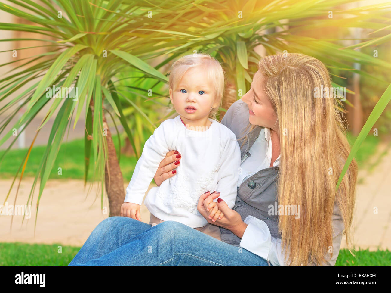 Felice madre con bambino ragazza seduta sul campo verde nel parco tropicale in una luminosa giornata di sole godendo della maternità, di amore e di felicità Foto Stock