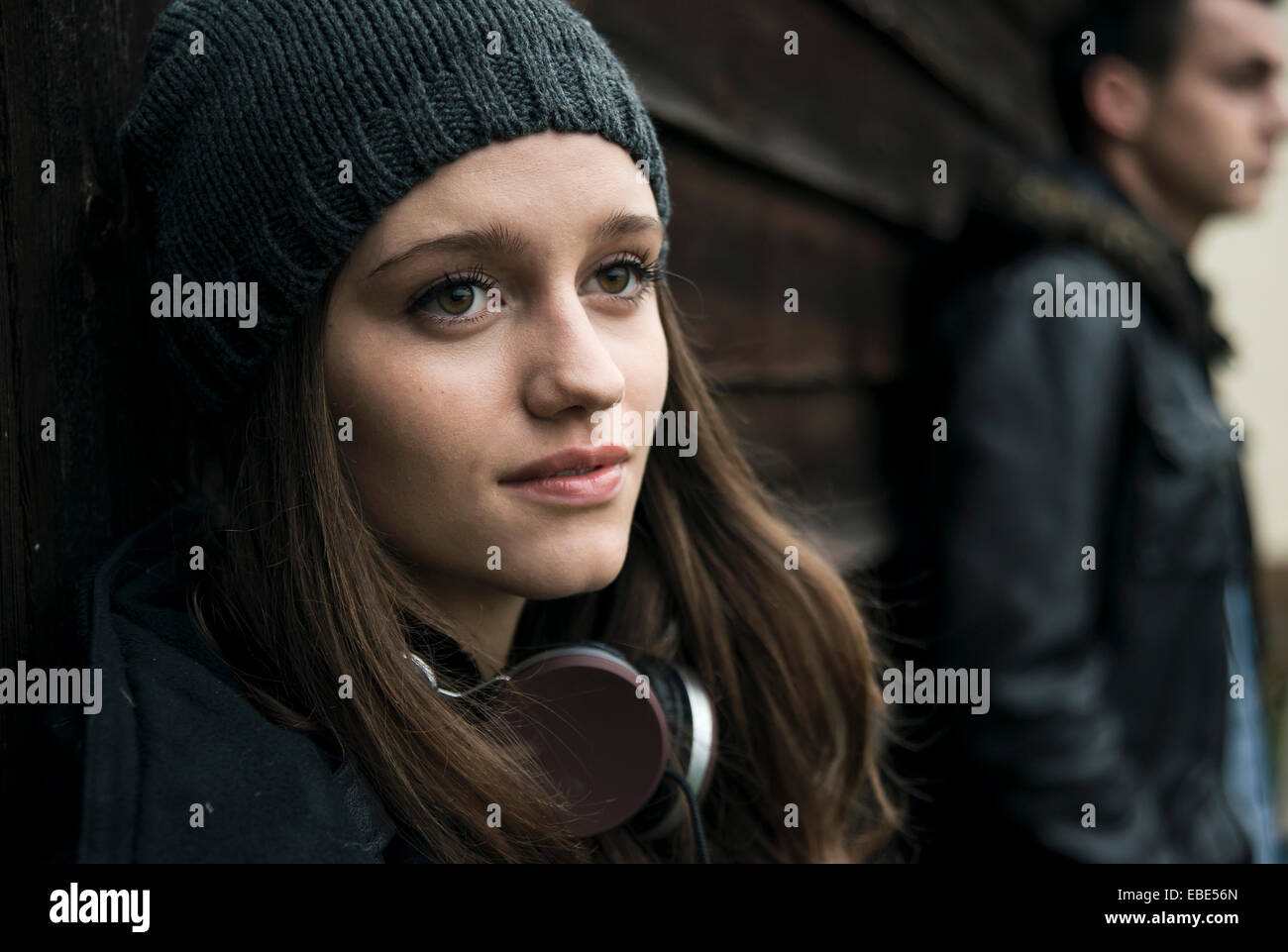 Close-up ritratto di ragazza adolescente all'aperto, indossare un cappello e cuffie intorno al collo con un giovane uomo in background, Germania Foto Stock