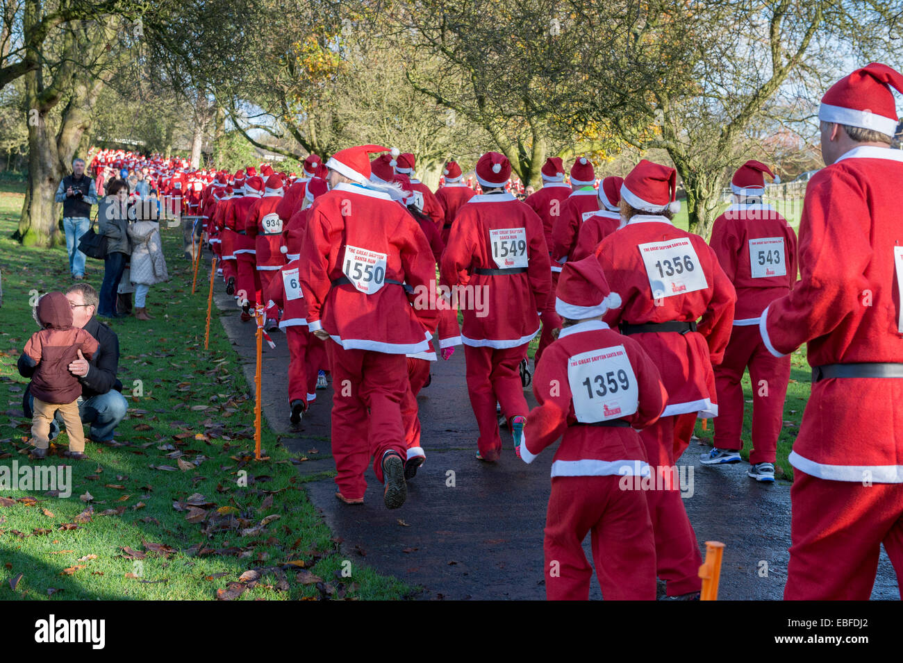 Vista posteriore di persone (uomini, donne e bambini) vestita in rosso e bianco di Babbo Natale abiti passeggiate, jogging, in esecuzione passato spettatori & prendendo parte alla grande Skipton Santa Fun Run, una raccolta di fondi annuale gara di beneficenza organizzata dal Rotary Club - Aireville Park, Skipton Town Center, North Yorkshire, Inghilterra, Regno Unito. Foto Stock