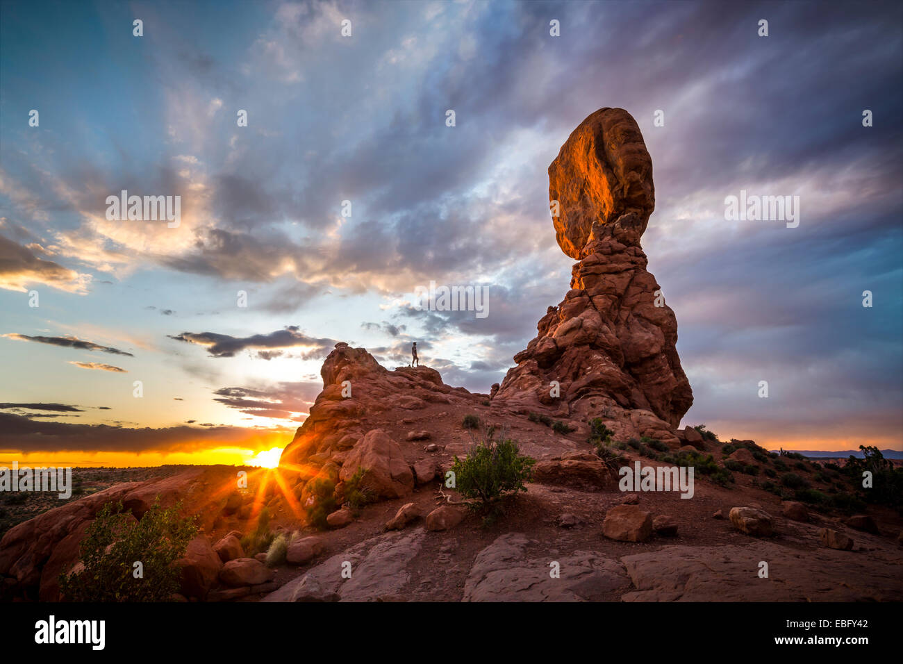 Whit Richardson in piedi sotto la roccia equilibrato nel Parco Nazionale di Arches, Moab, Utah. Foto Stock