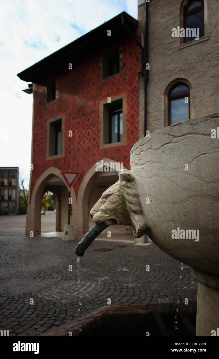 Fontana con testa di leone di fronte ad un edificio gotico con facciata decorata pieno di affreschi. Città vecchia di Pordenone. L'Italia. Foto Stock