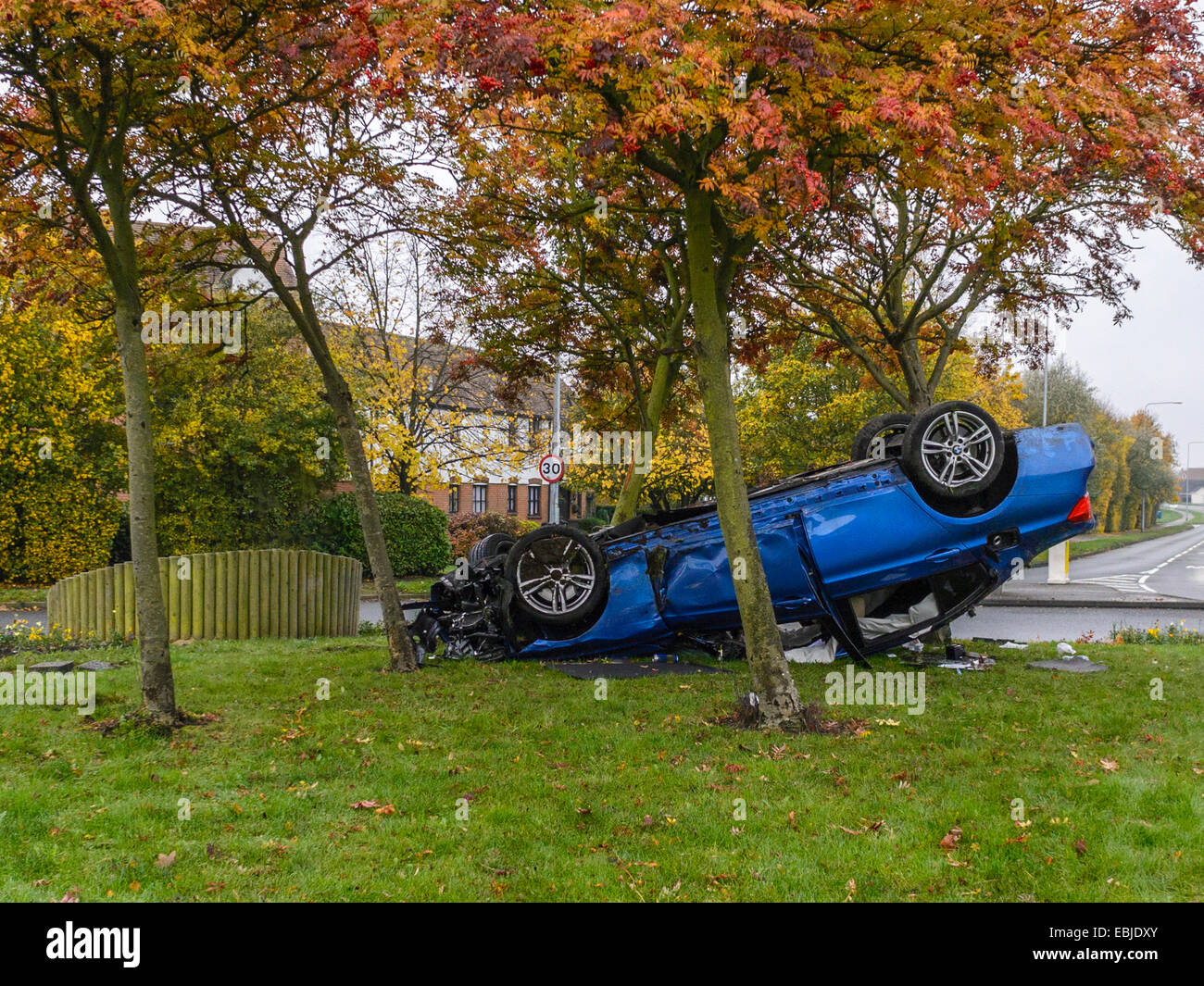 Insolito incidente automobilistico, raffiguranti blu berlina sportiva veicolo sul suo tetto, tra gli alberi su una rotatoria isola Foto Stock