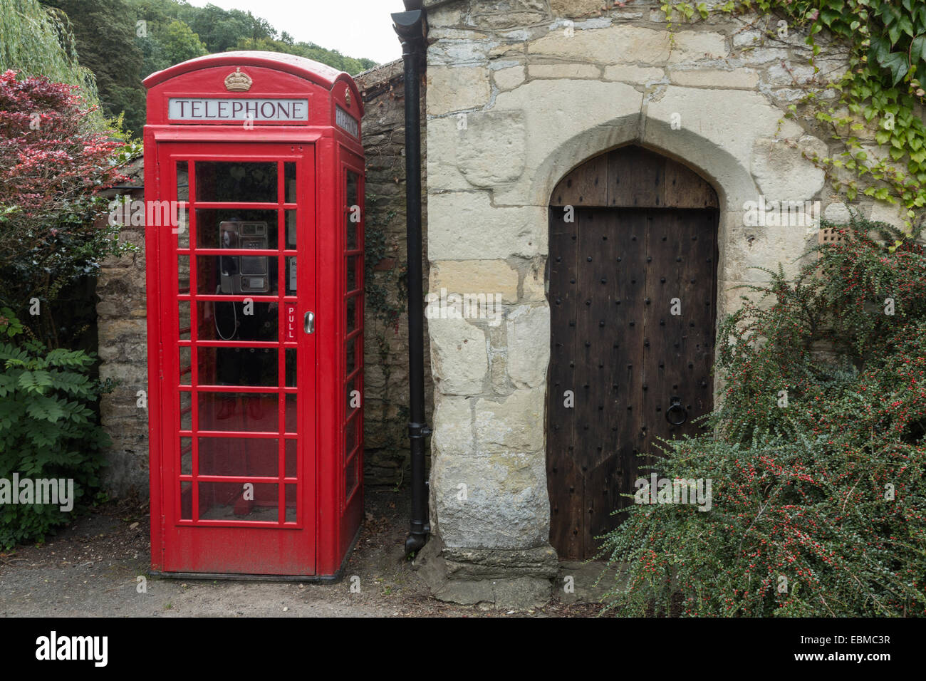 Telefono rosso casella accanto ad un vecchio legno chiodati porta in una cotswold edificio in pietra, Castle Combe, Wiltshire, Inghilterra Foto Stock