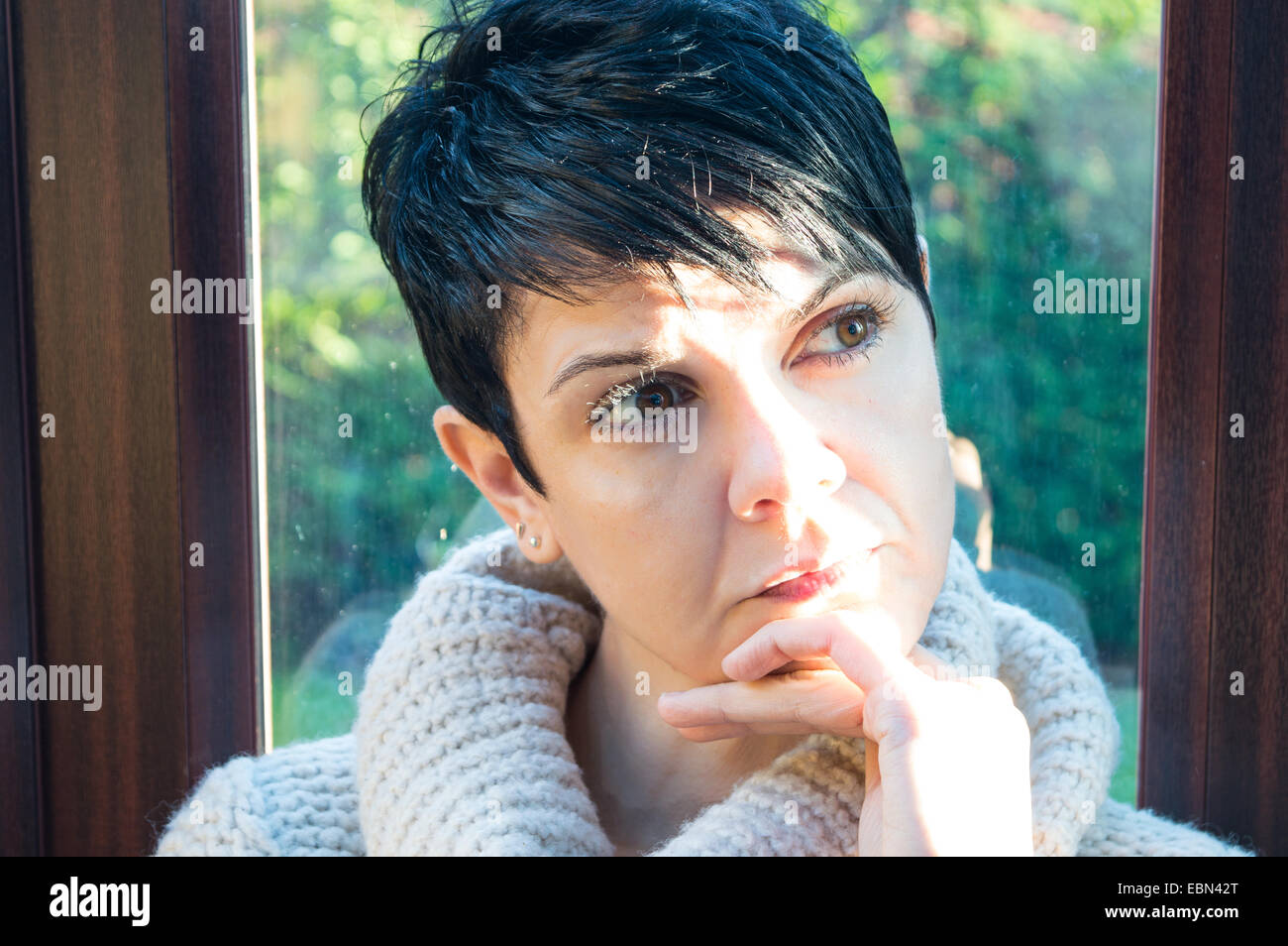 Bella giovane donna con breve, capelli neri guardando fuori da una finestra nel profondo pensiero con giardino in background e porta di patio Foto Stock