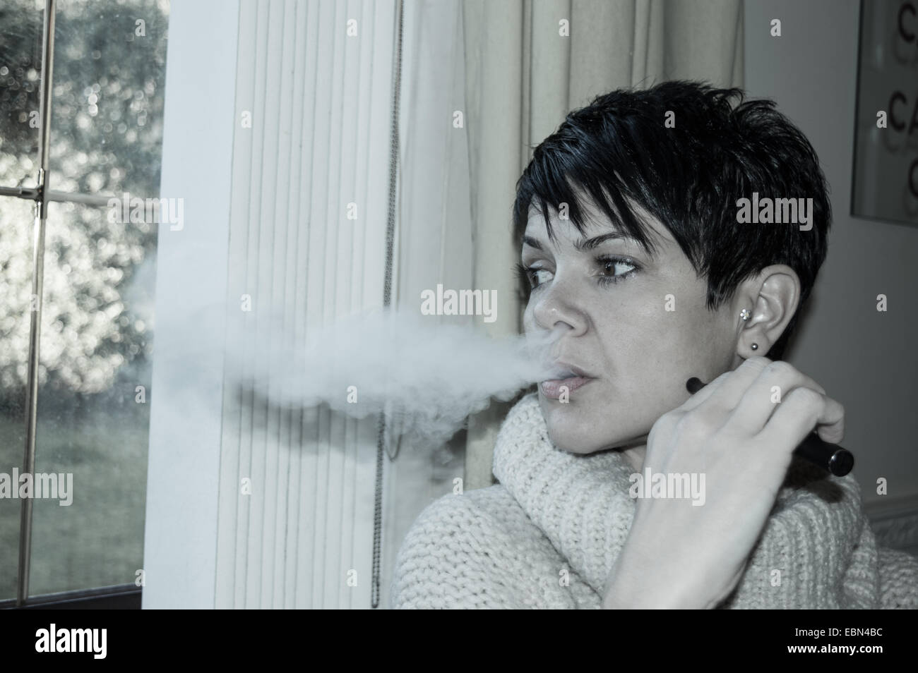 Bella breve, dai capelli scuri donna vaping e guardando fuori da una finestra nel profondo del pensiero Foto Stock